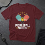 Maroon t-shirt with graphic of pickleball paddles, ball, and sun, text says "PICKLEBALL VIBES".