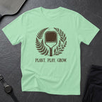 Mint green t-shirt with "PLANT. PLAY. GROW." and a pickleball paddle surrounded by foliage.