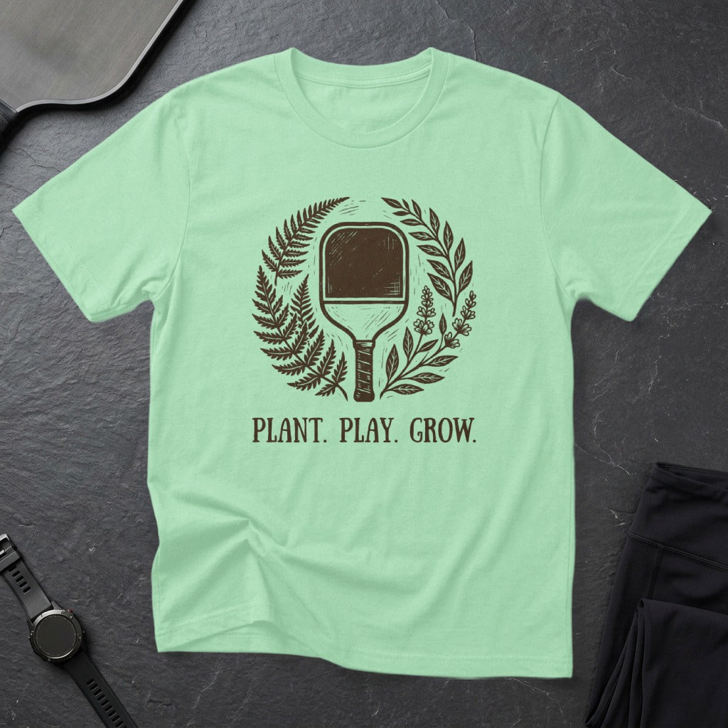 Mint green t-shirt with "PLANT. PLAY. GROW." and a pickleball paddle surrounded by foliage.