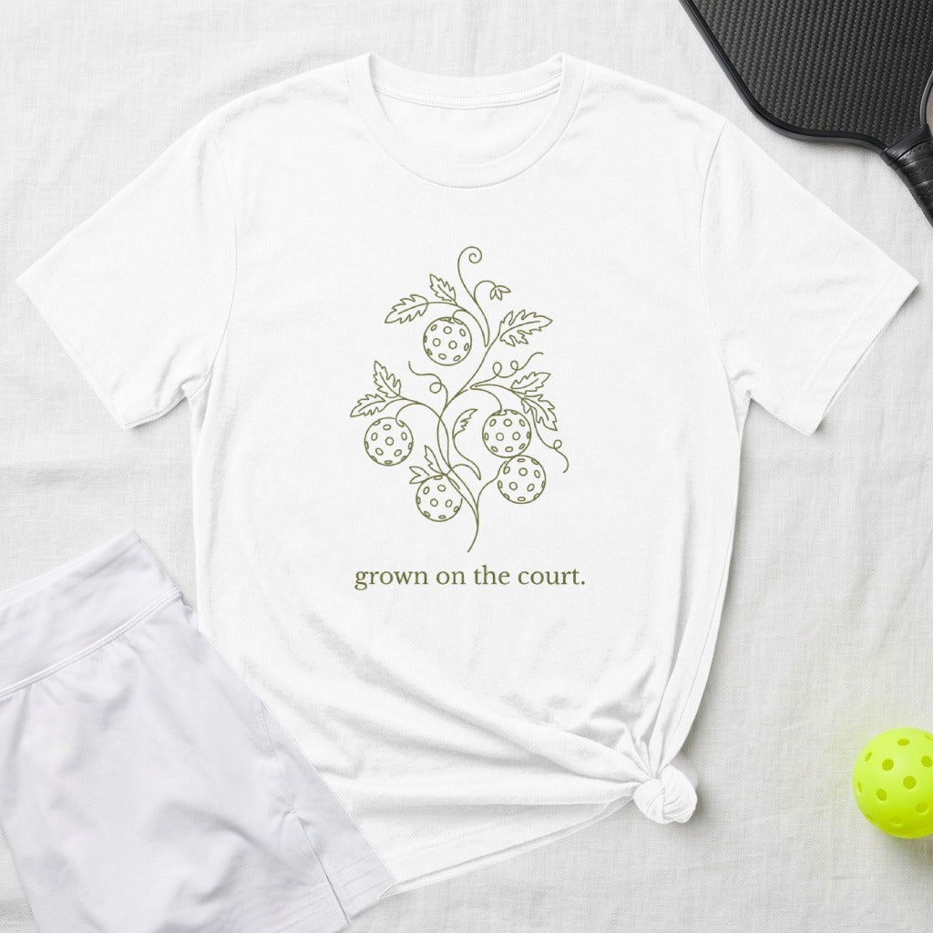 White t-shirt with a line drawing of pickleball balls on a vine and text "grown on the court."